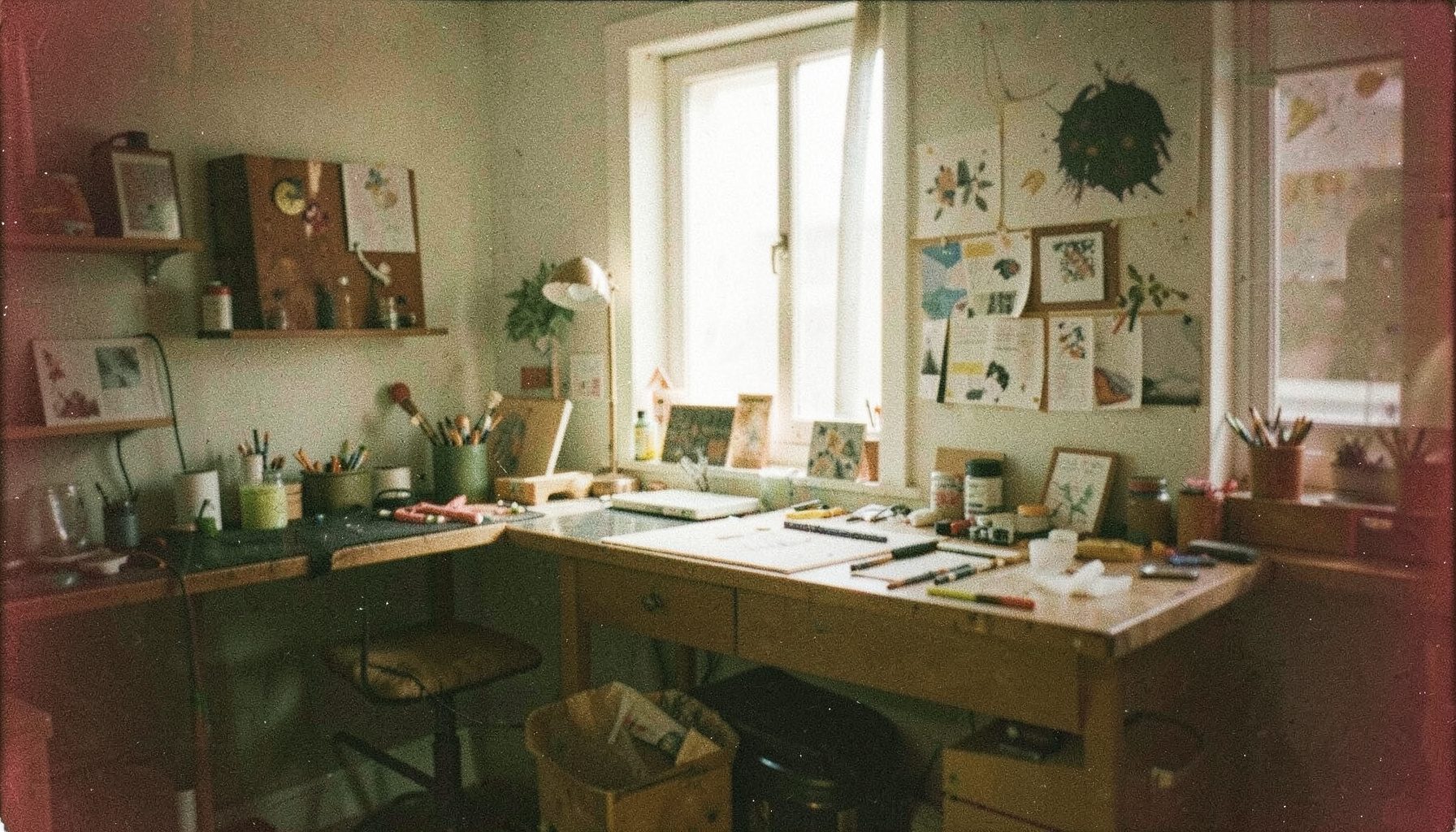 Warm-toned analog film photograph with visible grain showing a creative studio workspace with soft natural light, muted greens and earthy browns