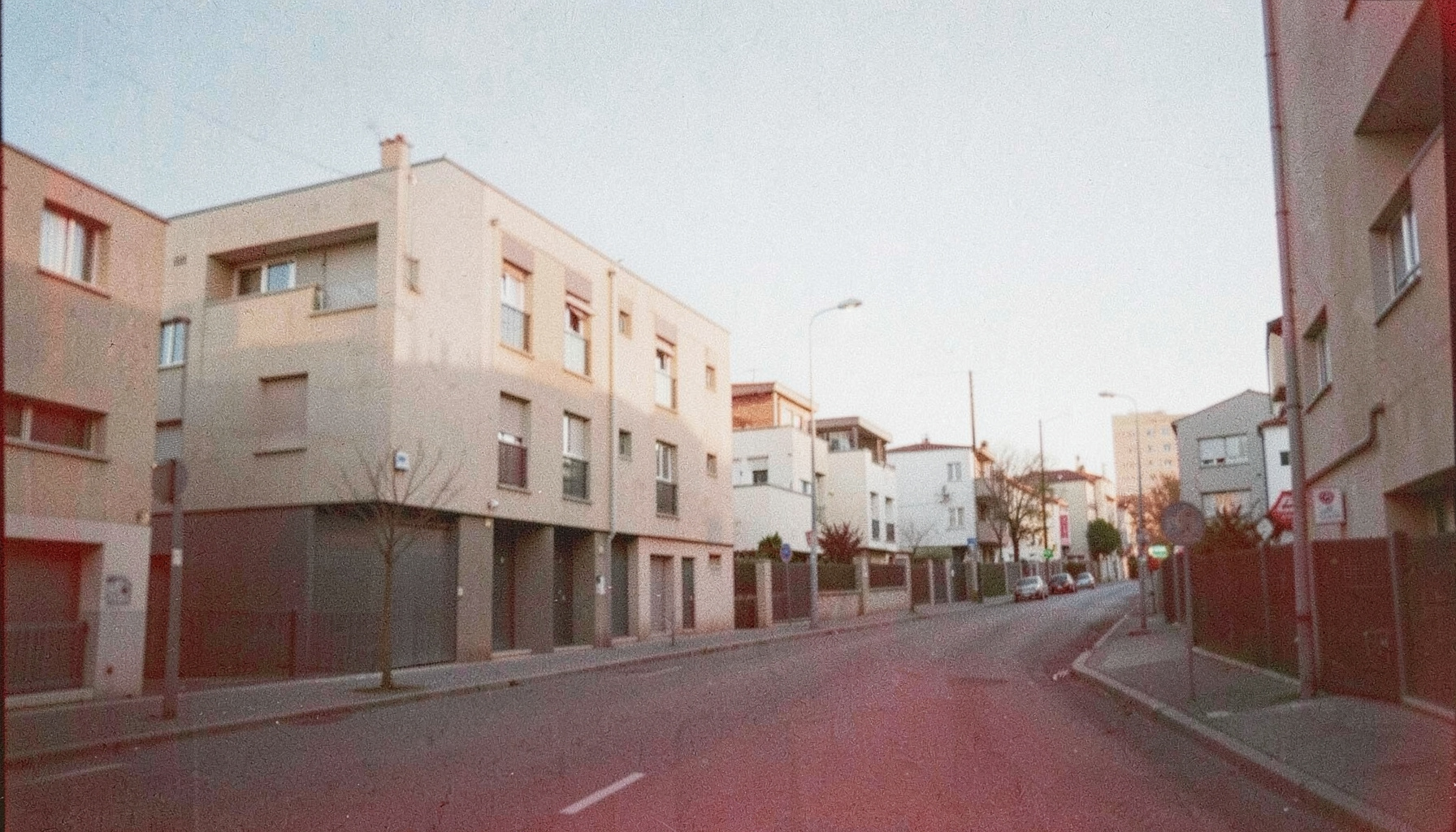 A grainy analog photograph of a quiet urban street in Villeurbanne near Lyon, soft morning light on modern low-rise buildings, muted beige and grey tones with pale blue sky