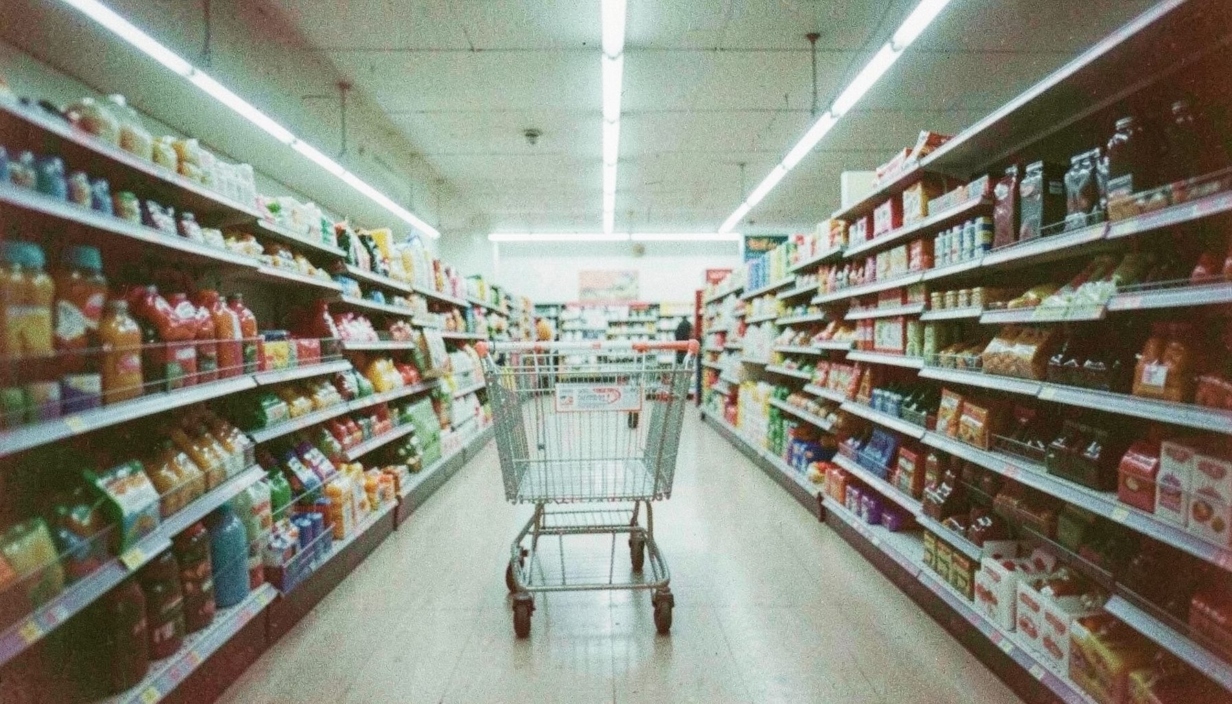 A supermarket aisle in harsh fluorescent light with a lone shopping cart, grainy analog photograph in cool whites and pale greens