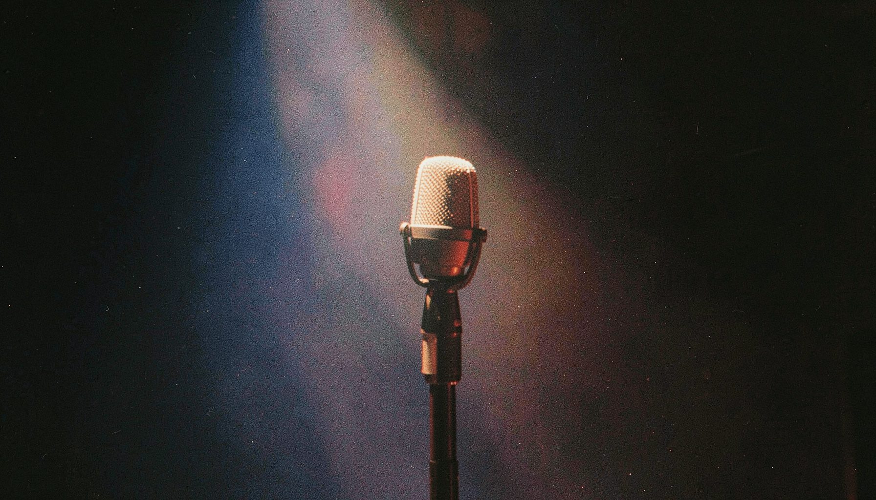 A microphone on a stand in a dimly lit room with a single spotlight, grainy analog film in deep blues and warm amber