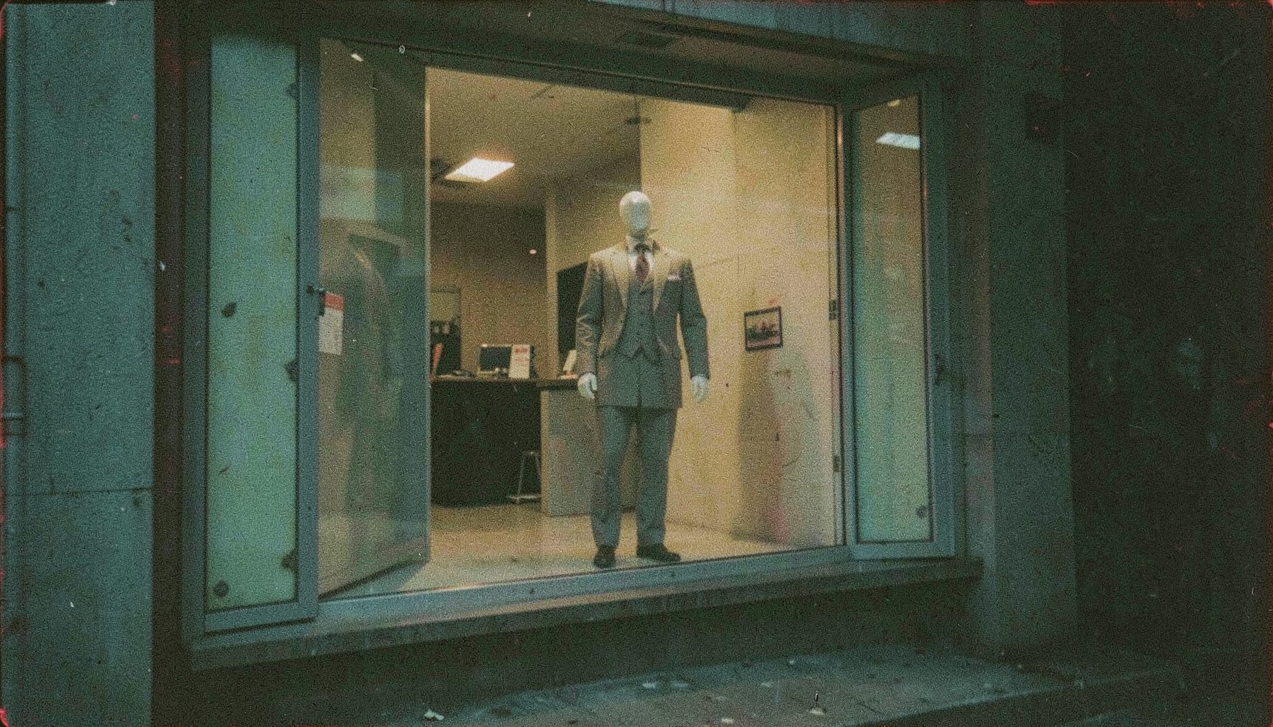 A mannequin standing in an empty storefront window at dusk, grainy analog photograph with muted teal and amber tones