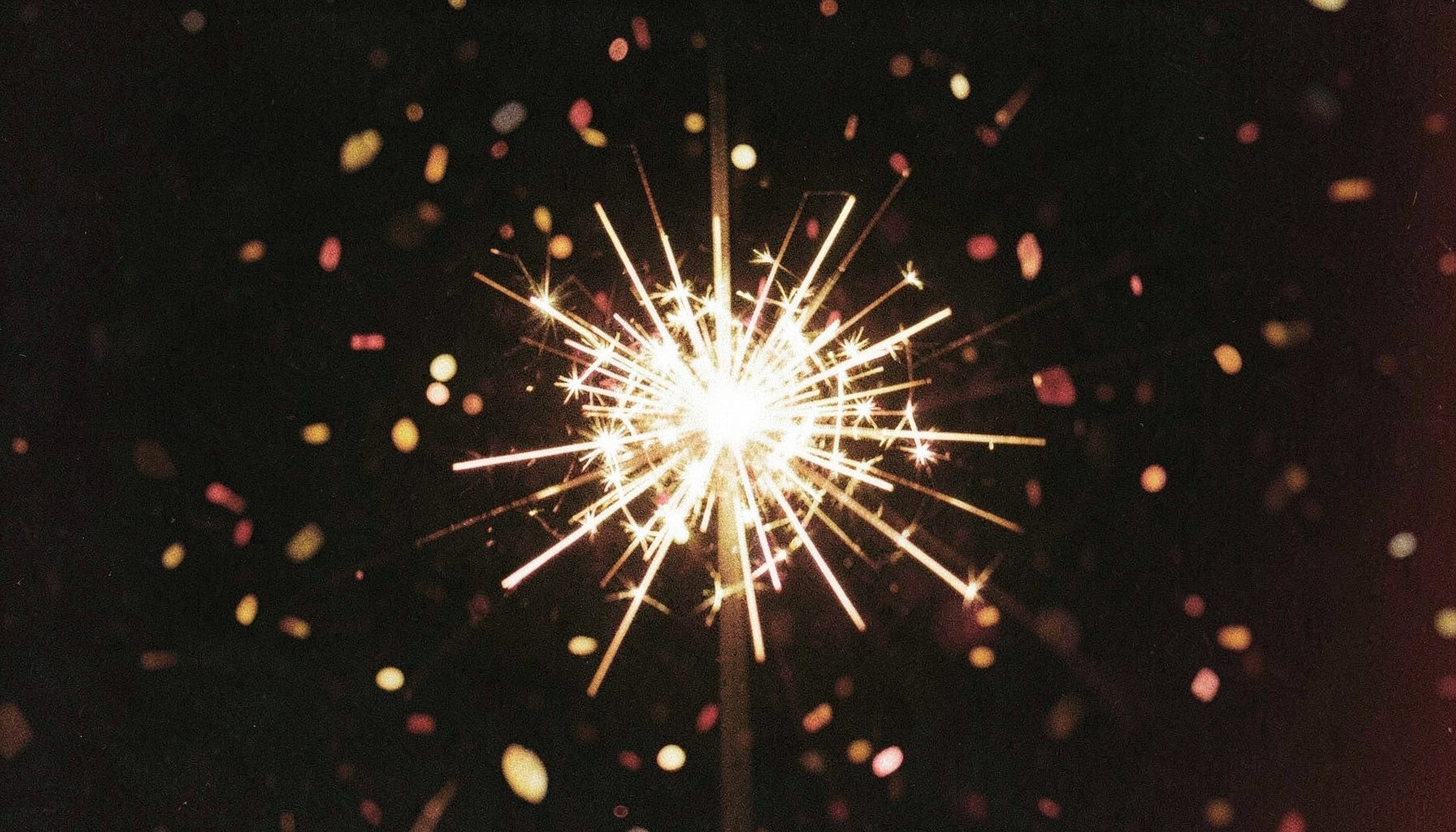 A festive close-up of sparklers and confetti against a dark background, grainy analog film in warm golds and deep blacks