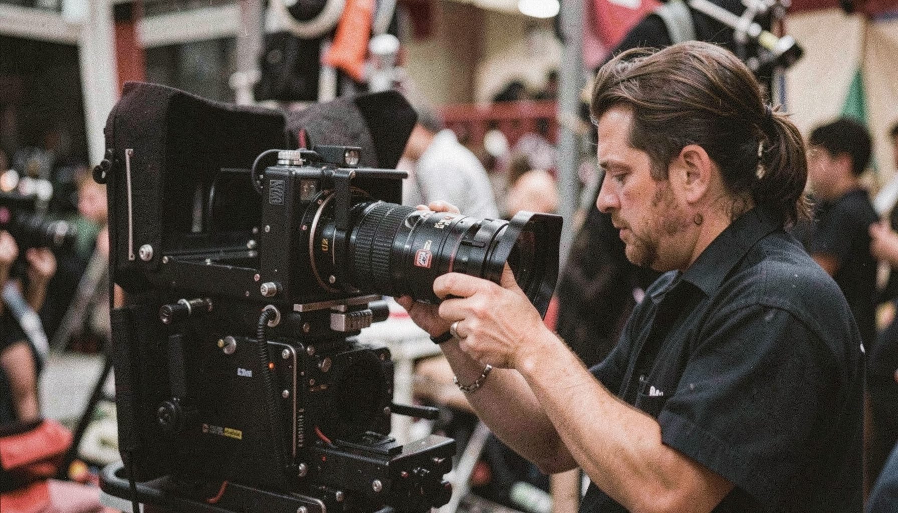 A camera assistant adjusting a large cinema lens on a matte box, focus-pulling rig visible, set against a blurred background of grip equipment; analog film photograph with visible grain, deep blacks and metallic silver highlights