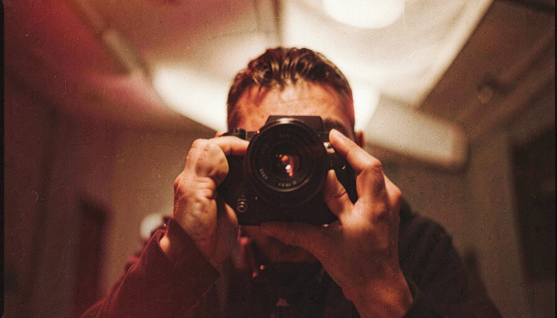 A director of photography looking through a viewfinder mounted on a cinema camera, framed by soft overhead diffusion; analog film photograph with visible grain, warm tungsten glow and deep shadow contrast