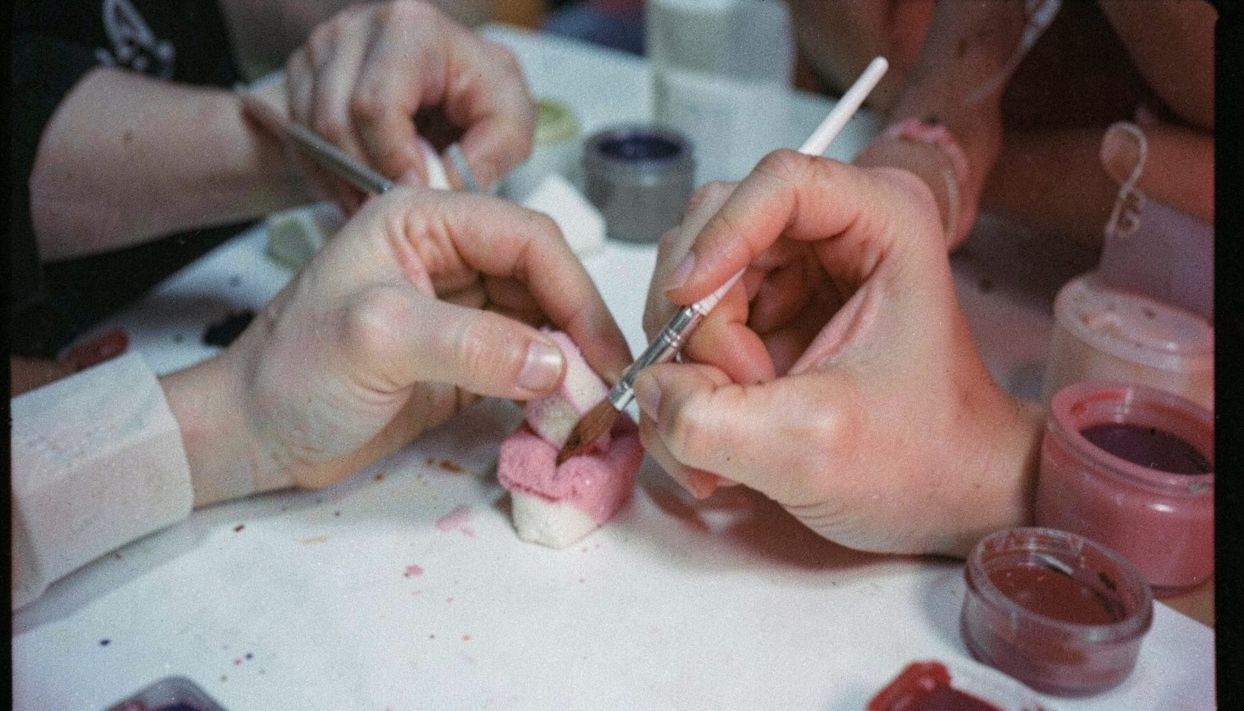 A close-up of hands applying prosthetic makeup with brushes and sponges on a pale surface, surrounded by small pots of pigment; analog film photograph with visible grain, muted skin tones and cool grey shadows