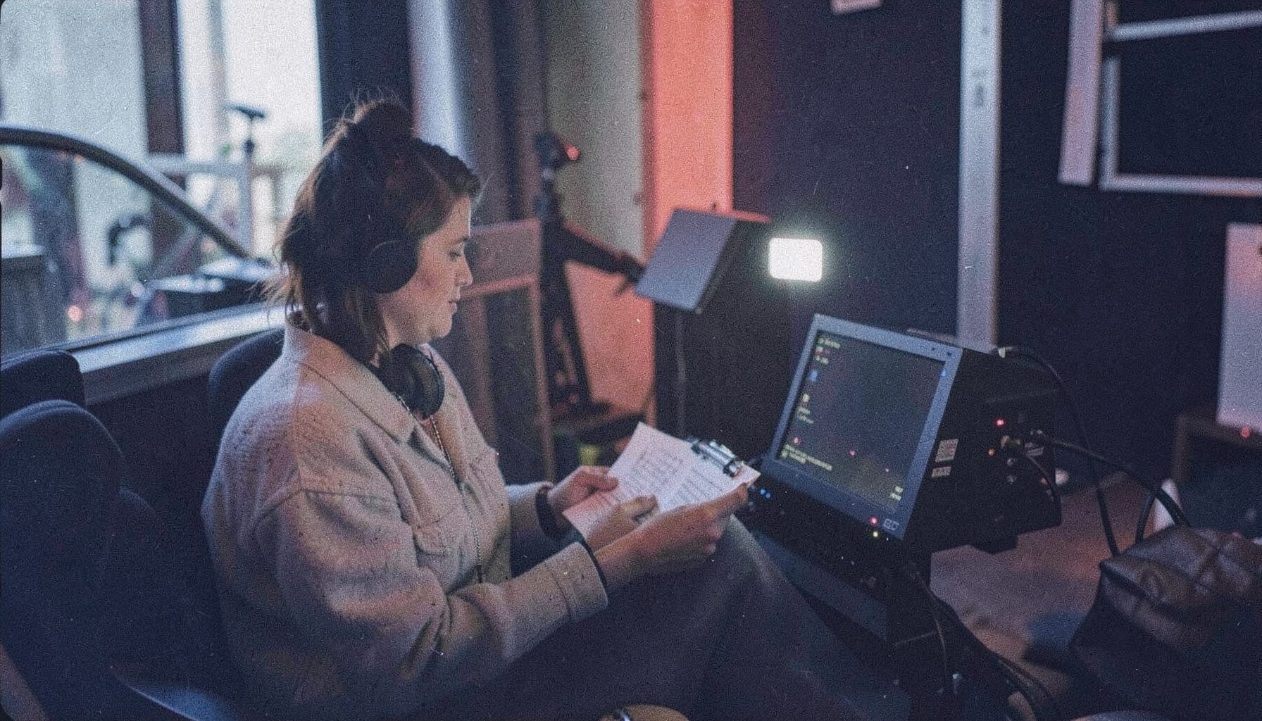 A woman seated at a small monitor on a film set, holding a clipboard and wearing headphones, with script pages visible in her lap; analog film photograph with visible grain, soft natural light and muted navy tones